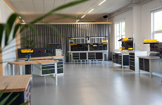 Der RAU Ausstellungsraum mit mehreren Werkbänken, Werkzeugaufbewahrung und hellen Fenstern, umgeben von grauen Wänden und einer modernen, gut organisierten Atmosphäre. Eine Pflanze befindet sich im Vordergrund.
