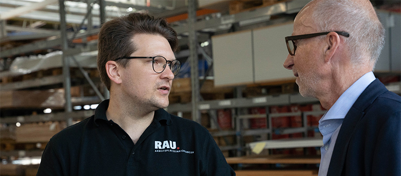 Zwei Männer stehen in einem Lagerhaus und unterhalten sich. Der linke Mann trägt ein schwarzes Polohemd mit der Aufschrift "RAU Arbeitsplatzeinrichtungen". Regale und Produkte sind im Hintergrund.