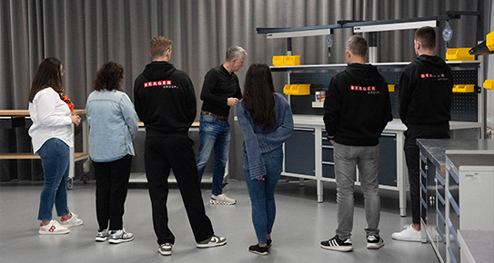 Personengruppe steht in einem Raum, hört einem präsentierenden Mann zu. Der Raum enthält Arbeitsplätze mit Regalen und gelben Behältern. Personen tragen schwarze Hoodies mit rotem Logo.