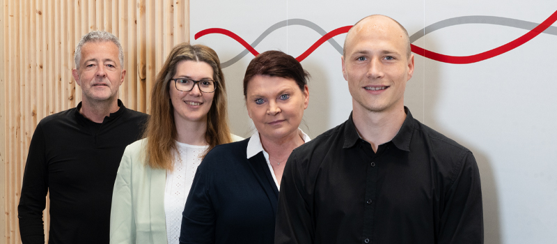Vier Personen stehen nebeneinander, lächeln leicht, vor einer Wand mit Holzverkleidung und geschwungenen Linien. Alle tragen formelle Kleidung.