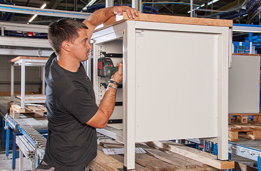 Ein Mann montiert ein weißes Metallgehäuse mit einer Bohrmaschine in einer Fabrikhalle
