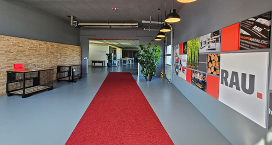 Roter Teppich führt durch modernen Eingangsbereich mit Werbetafeln, Pflanzen und Holzakzenten, dekoriert mit Hängelampen und Metallstrukturen. Text enthält: „RAU“.