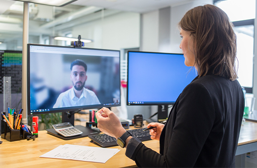 Frau führt mit einem Mann auf dem Bildschirm ein Online-Meeting in einem modernen Büro mit Schreibtisch, Dokumenten, Stiften und zwei Monitoren.