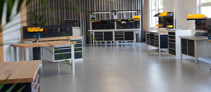 Werkbänke stehen in einem hellen Raum mit grauem Boden, umgeben von Werkzeugwänden und Stauraum. Pflanzen und Fenster verstärken die offene, geordnete Atmosphäre.