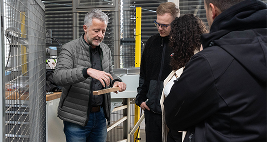 Ein Mann zeigt einer Gruppe von vier Personen ein Brett in einer industriellen Umgebung mit Regalen und Metallzaun.